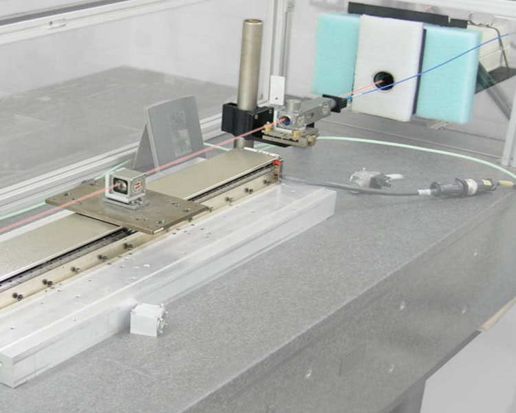 A precision measurement device on a workbench consisting of a long metallic rail, digital sensors, a cable, and a vertical cylindrical component adjacent to a foam-padded piece in the background.