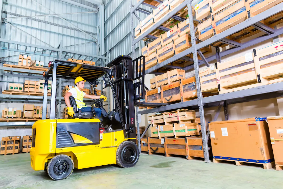 A worker operates a yellow forklift in a warehouse, moving wooden crates on a metal shelf. The worker wears a hard hat and a high-visibility vest.