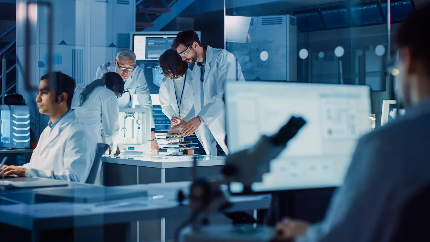 A group of scientists wearing lab coats work collaboratively in a modern laboratory, surrounded by various scientific equipment and computer monitors.