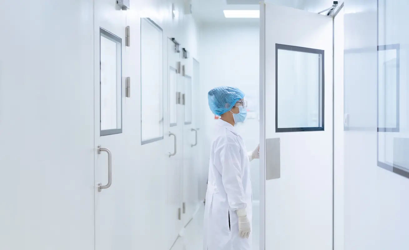 A person wearing protective clothing, including a hair cover, mask, gloves, and lab coat, opens a door in a cleanroom environment.