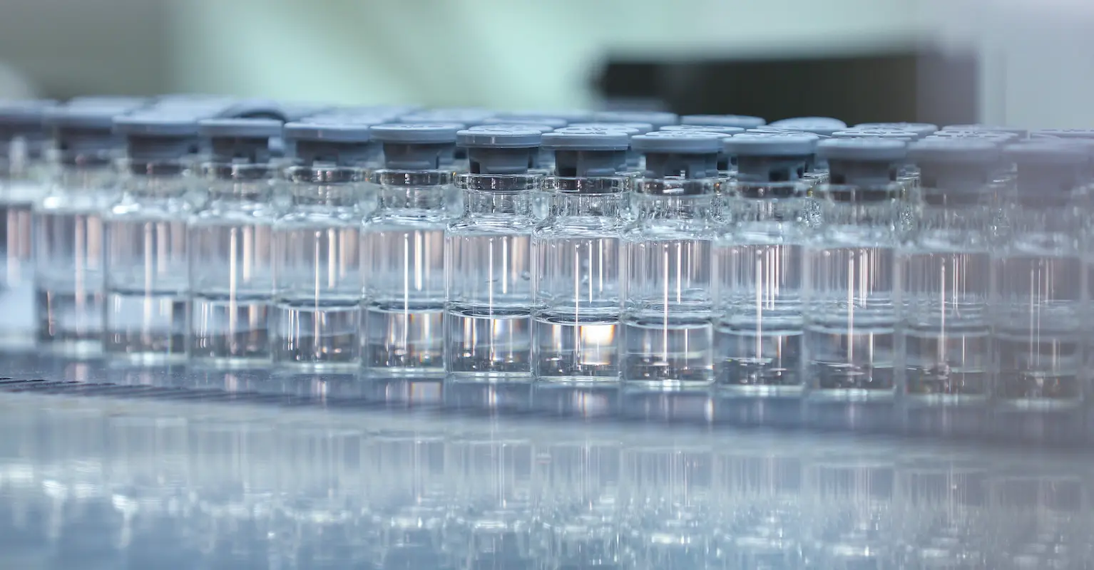 Rows of glass vials with gray caps, filled with a clear liquid, are aligned on a reflective surface in a laboratory setting.