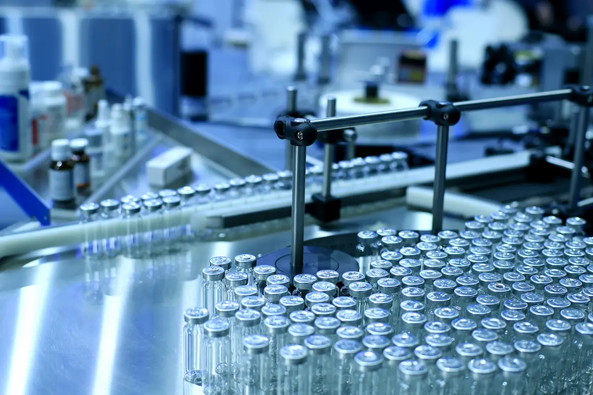 Image of a pharmaceutical production line with numerous glass vials being processed and various equipment used for manufacturing in a sterile industrial environment.