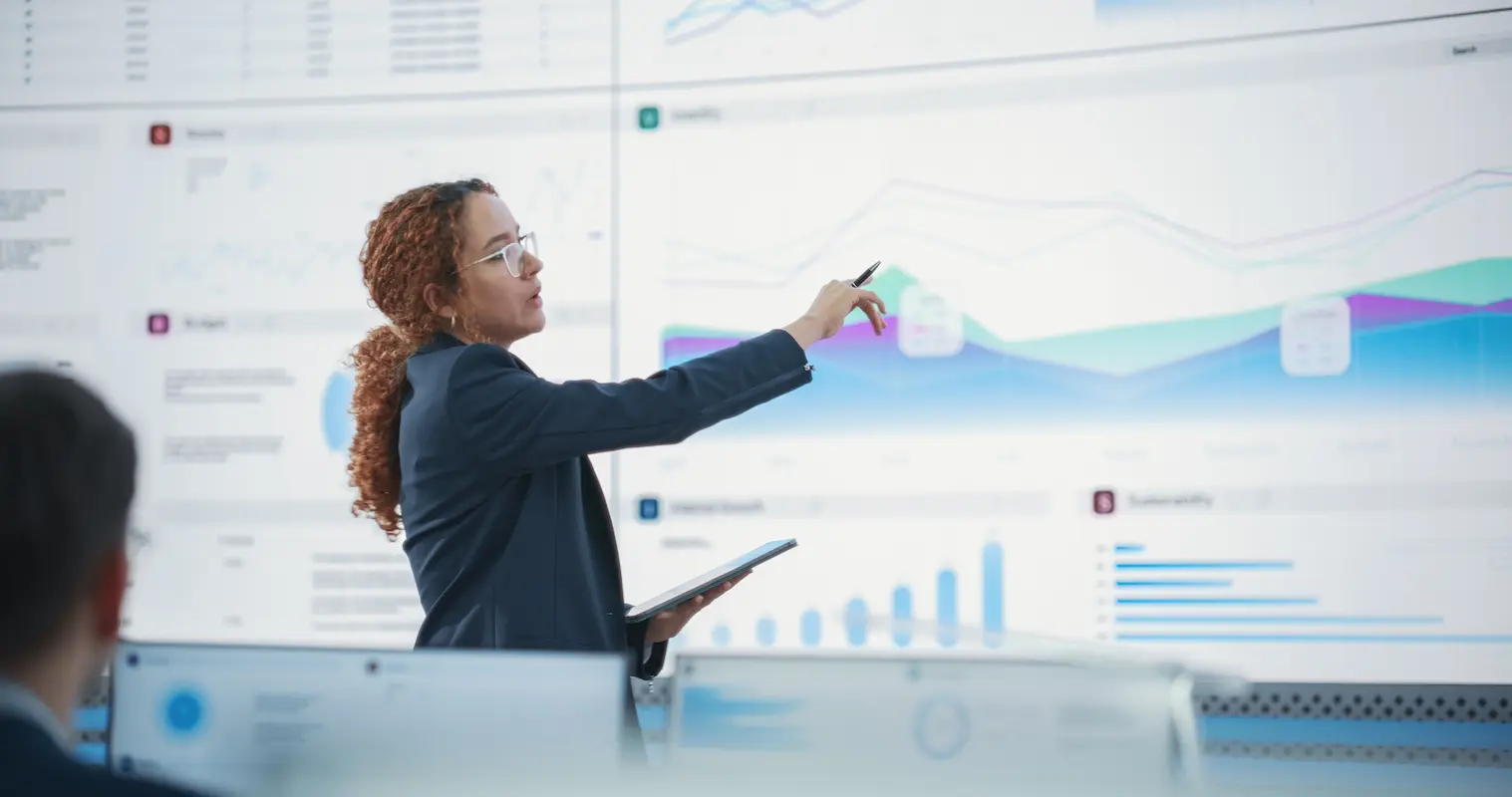 A person in business attire stands in front of large digital screens displaying graphs and charts, pointing at the data while holding a tablet.