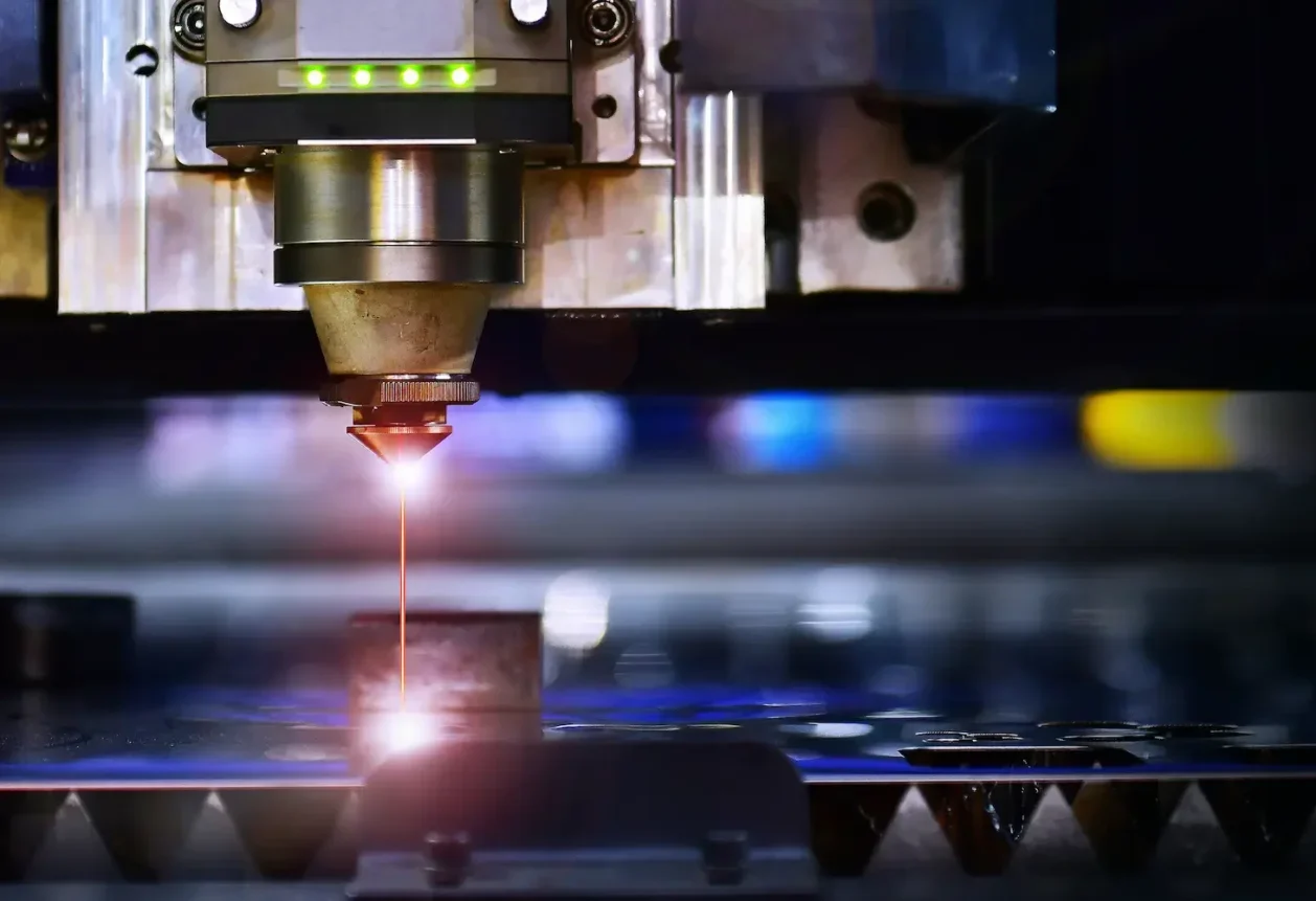 A laser cutting machine processes a piece of metal, with a focused beam and precise cutting action visible.