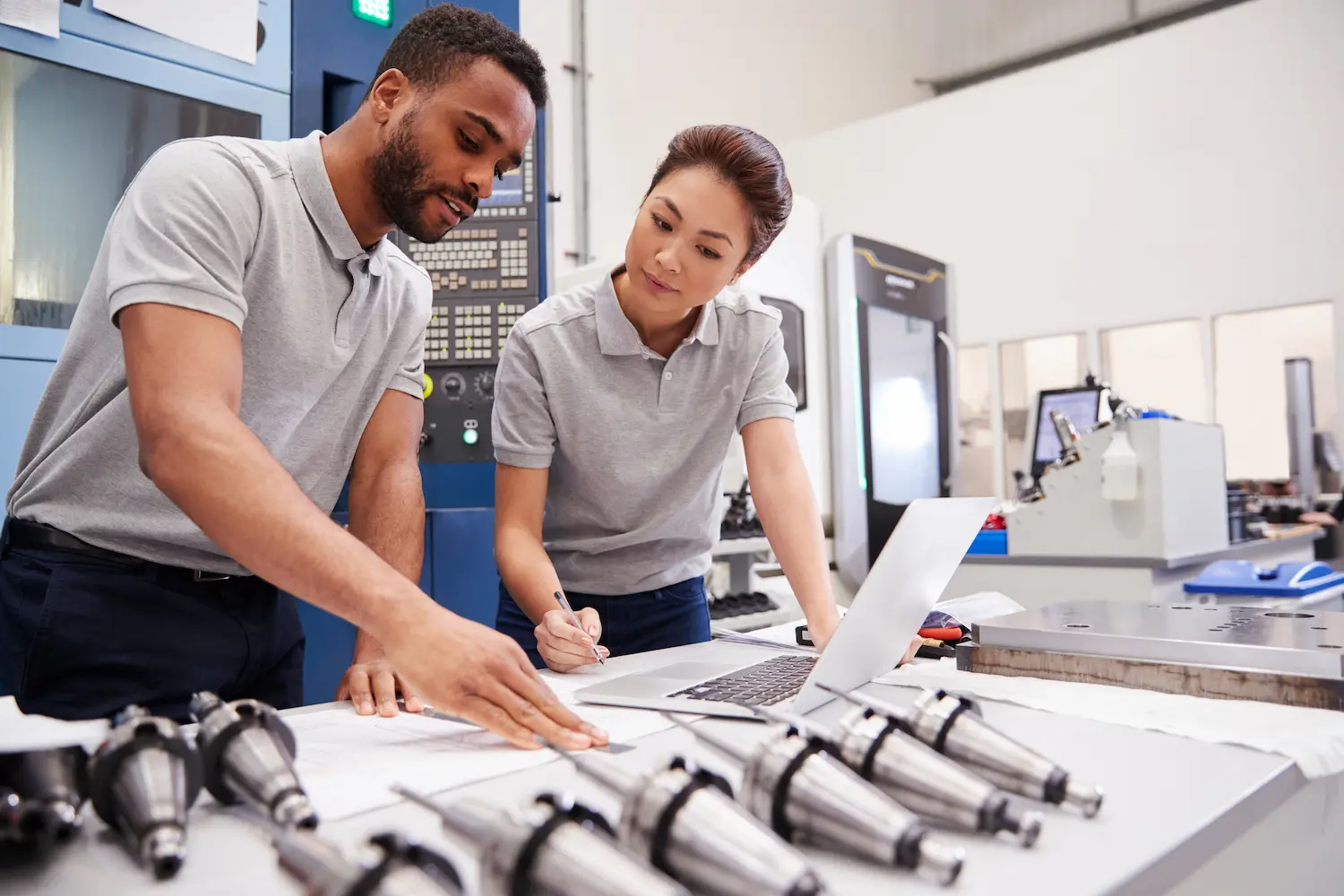 Two industrial workers, wearing grey polos, discuss over documents and a laptop in a factory setting. Multiple machine parts are arranged on the table beside them.