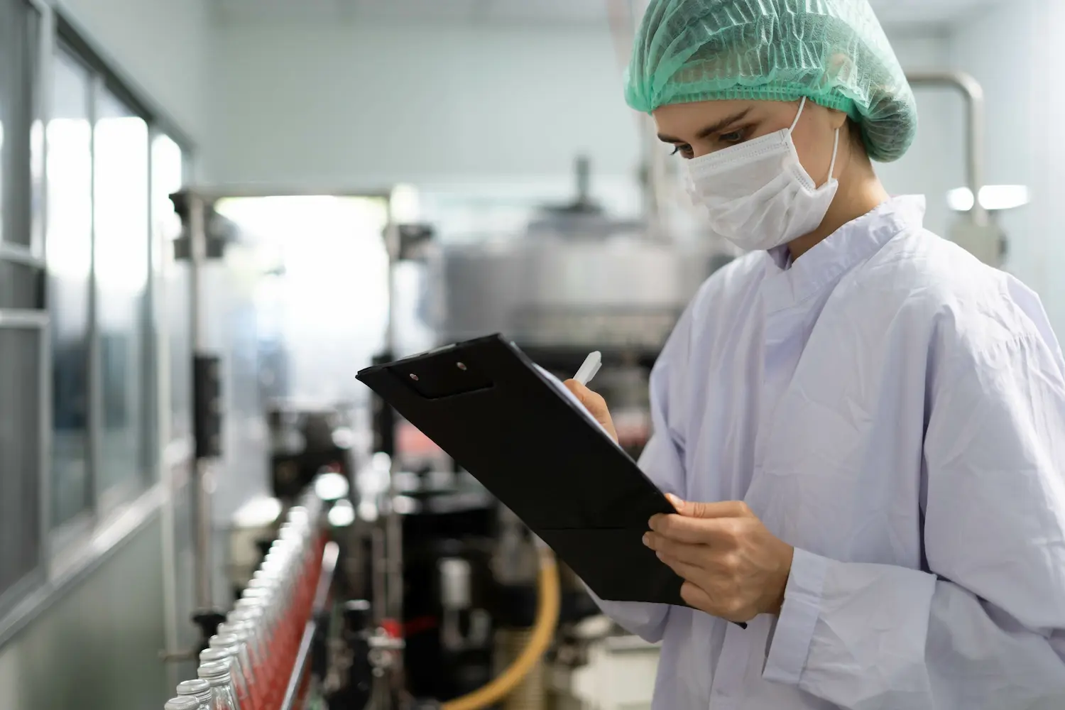 A person in a lab coat and hair net, wearing a face mask, writes on a clipboard in a laboratory filled with equipment and rows of sample bottles.