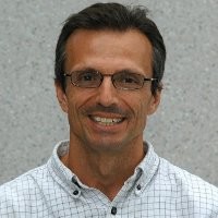 A man wearing glasses and a light-colored, checkered shirt smiles against a neutral background.