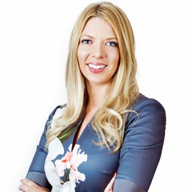 Blonde woman with long hair smiles while standing with arms crossed, wearing a gray top with a floral design.