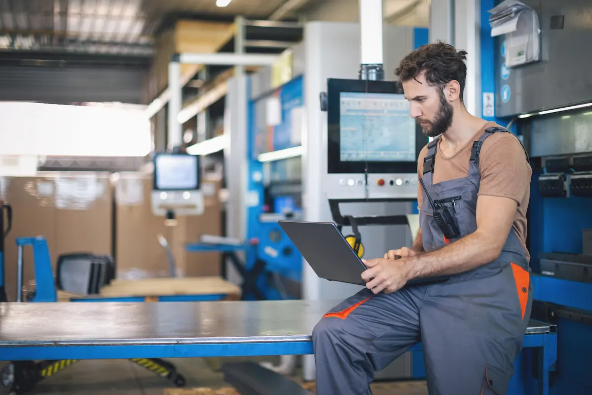 A man in work overalls uses a laptop in an industrial setting, with machinery and equipment in the background.