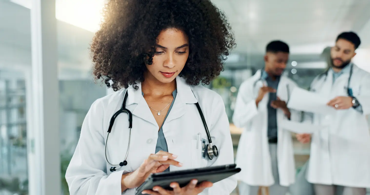 A doctor in a white coat uses a tablet in a hospital hallway. Two other doctors are talking in the background.