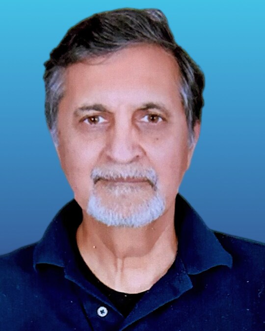 A man with short, graying hair and a beard against a blue background, wearing a dark collared shirt.