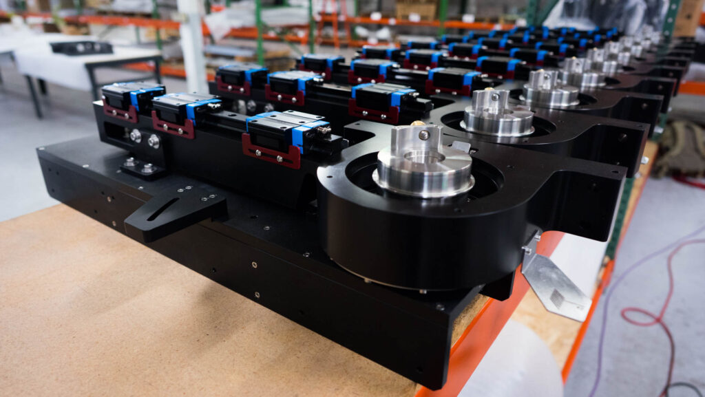 A row of black mechanical components with metal fixtures and blue and red parts is aligned on a wooden table in an industrial setting.