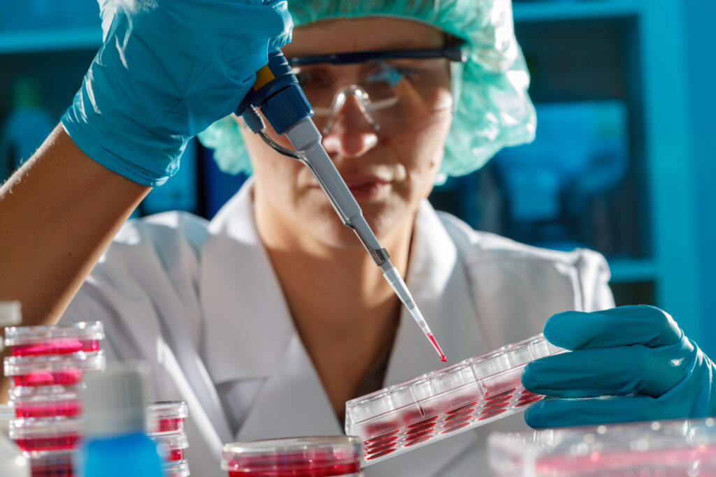 Scientist in lab coat and gloves uses a pipette to transfer liquid into a petri dish.